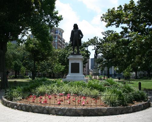 Bronzen standbeeld van een man tussen rode bloemen in een park in New Orleans met gebouwen op de achtergrond