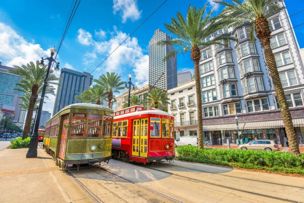 Een groene en een rode tram staan naast elkaar op straat, omgeven door palmbomen met wolkenkrabbers van New Orleans op de achtergrond