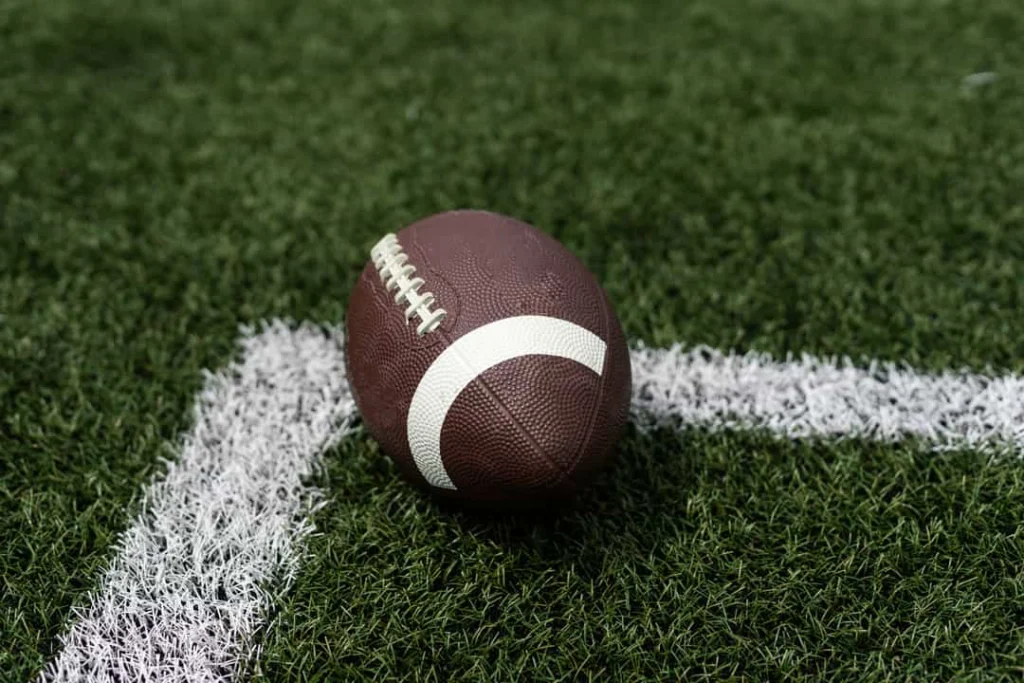 Een bruine american football met witte veters ligt op een kunstgrasveld bij de kruising van witte lijnen