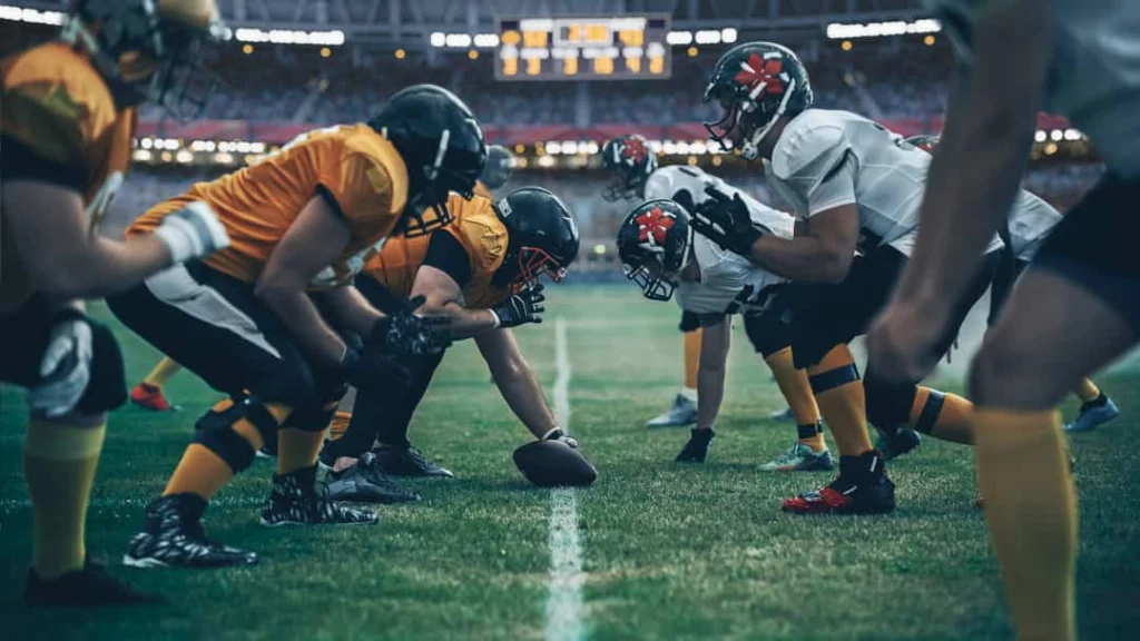 American football spelers in gele en witte shirts staan tegenover elkaar bij de scrimmagelijn in een verlicht stadion in New York