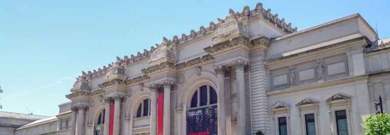 Klassieke stenen gevel met zuilen van het American Museum of Natural History in New York onder een blauwe hemel, met rode banieren aan de ingang