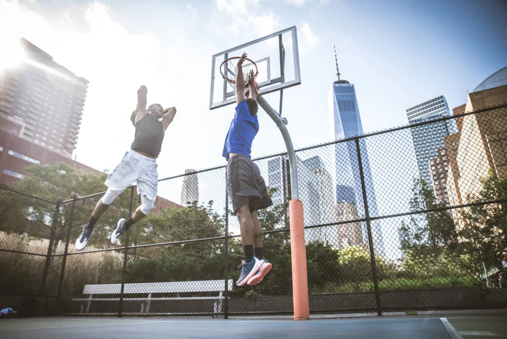Twee basketballers springen naar de ring op een omheind stadspleintje met wolkenkrabbers van New York op de achtergrond