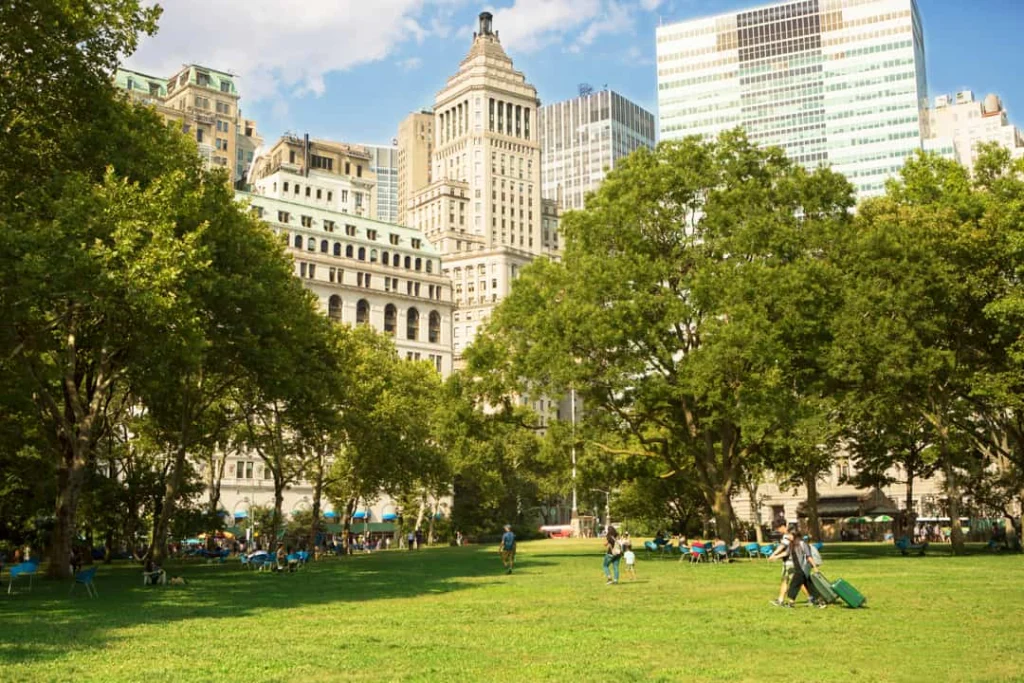 Groen grasveld in Battery Park New York met ontspannen bezoekers en bomen, met op de achtergrond de toren van de Manhattan Municipal Building