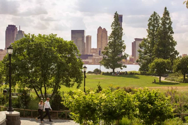 Wandelend stel op een pad in Battery Park New York met groene bomen en uitzicht op wolkenkrabbers aan de overkant van het water