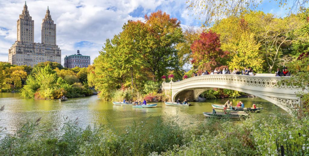 Roeibootjes varen onder de witte Bow Bridge in Central Park tussen bomen in oranje en rode herfsttinten met de San Remo torens op de achtergrond