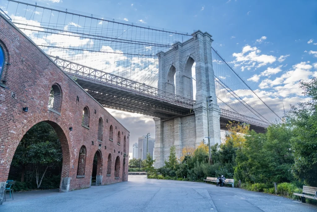 De Brooklyn Bridge met stenen pijlers en kabels gezien vanaf een plein met een oude bakstenen loods in de wijk Dumbo in New York onder een blauwe lucht