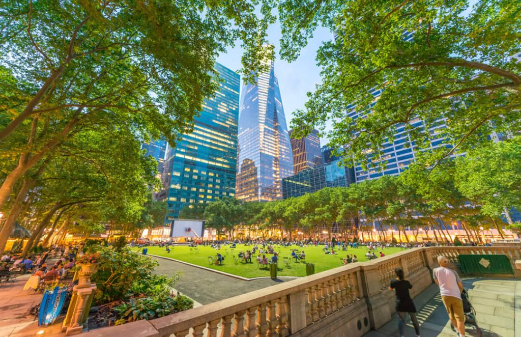 Groen grasveld van Bryant Park omgeven door bomen met bezoekers op het gras en verlichte wolkenkrabbers erachter bij schemering