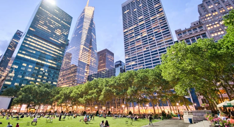 Groen grasveld in Bryant Park met bezoekers op stoelen tussen bomen terwijl verlichte wolkenkrabbers oprijzen tegen de avondlucht