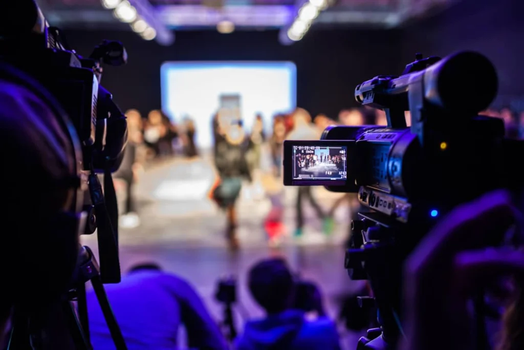 Twee videocamera's op de voorgrond filmen een model dat wazig op de achtergrond over de catwalk loopt