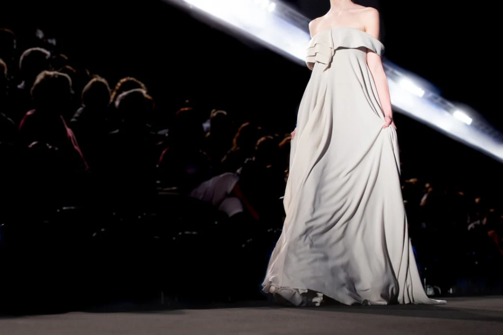 Model in een lange lichtgrijze avondjurk met blote schouders loopt over de catwalk met een spotlight op de achtergrond