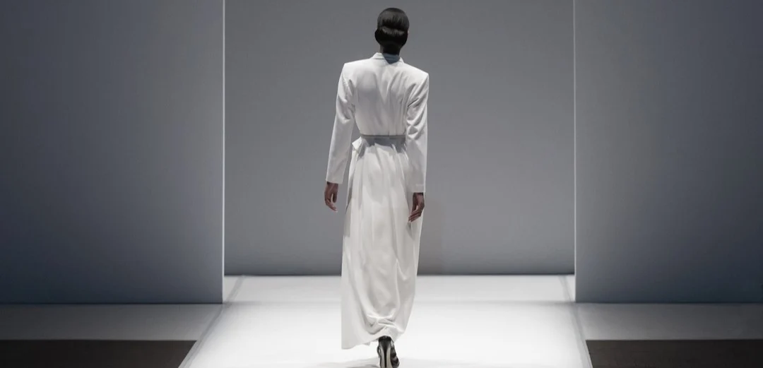 Model loopt in een lange witte jas met ceintuur over de catwalk tijdens een modeshow