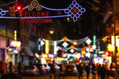 Wazig beeld van een verlichte boog met de tekst Welcome to Chinatown boven een drukke straat in New York in de avond met kleurrijke lichtjes en voetgangers