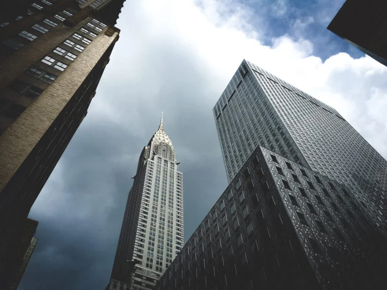 Aanzicht op de art deco top van de Chrysler Building die oprijst tussen donkere wolkenkrabbers onder een bewolkte lucht