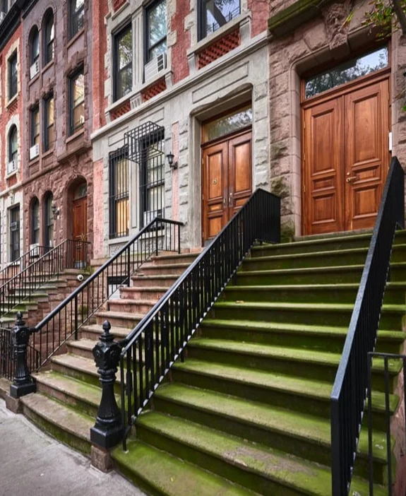 Bemoste stenen trap met zwarte leuningen naar de houten voordeuren van een rij bruinstenen woonhuizen in New York City