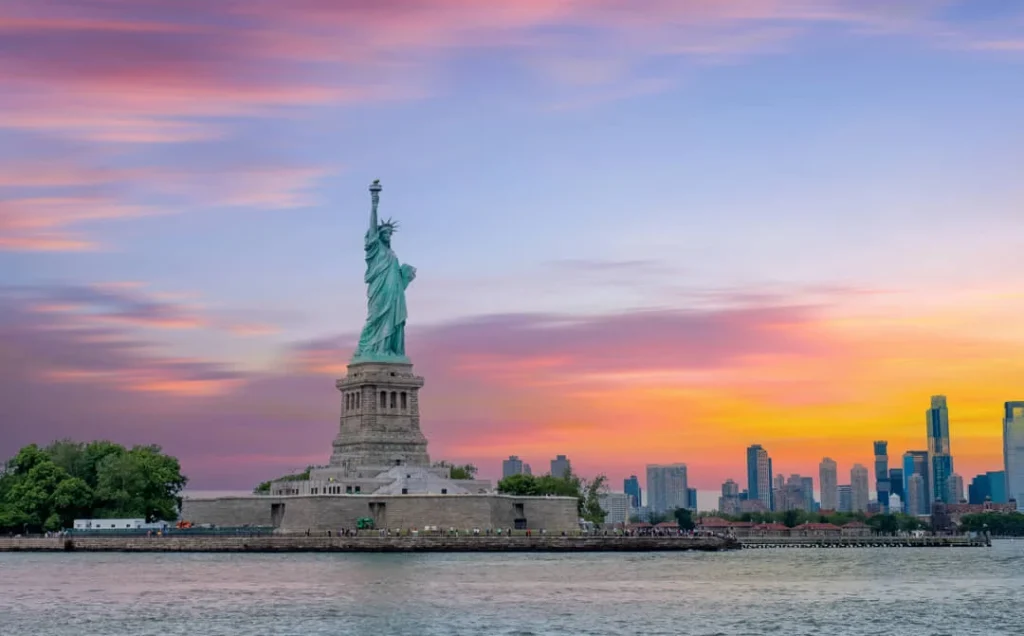 Het Vrijheidsbeeld silhouetteert tegen een roze en oranje avondlucht, met de skyline van New York City in de verte