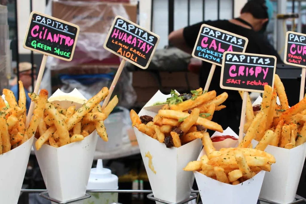 Close-up van meerdere bekertjes friet met kruiden en sauzen voor handgeschreven bordjes op een drukke streetfoodmarkt