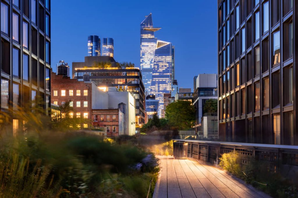 Verlicht houten wandelpad van de High Line tussen groen met de glazen wolkenkrabbers van Hudson Yards op de achtergrond in de avond