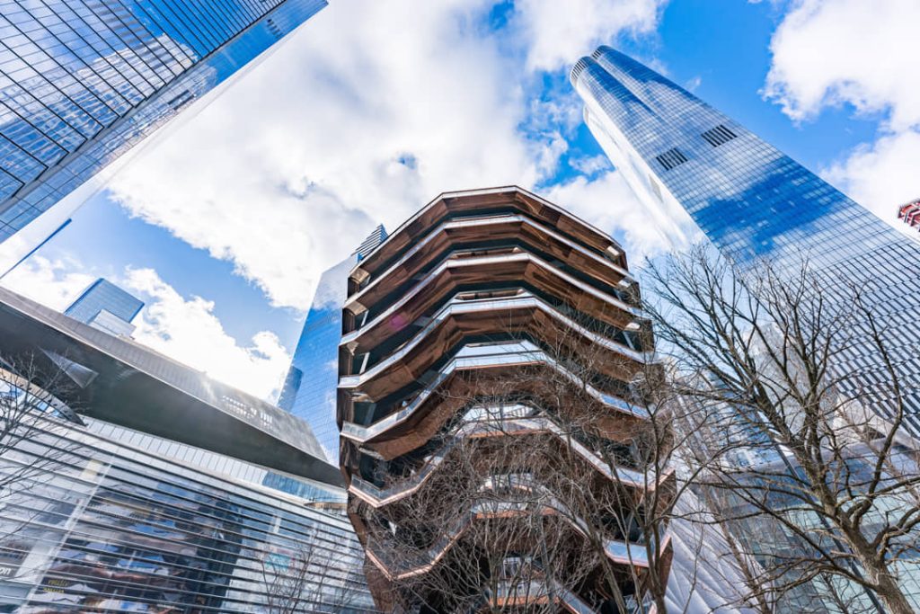 Blik recht omhoog naar de honingraatvormige trappenstructuur van de Vessel omringd door wolkenkrabbers bij Hudson Yards in New York