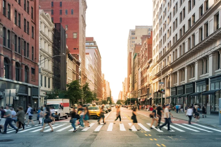 Voetgangers steken op een zebrapad over op Fifth Avenue tussen bakstenen en glazen gebouwen terwijl warm avondlicht de straat vult