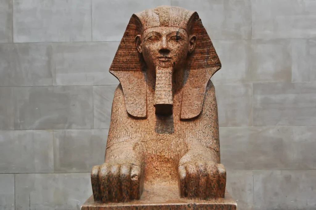 Een stenen sfinxbeeld met beschadigd gezicht tentoongesteld tegen een grijze stenen muur in het Metropolitan Museum of Art in New York