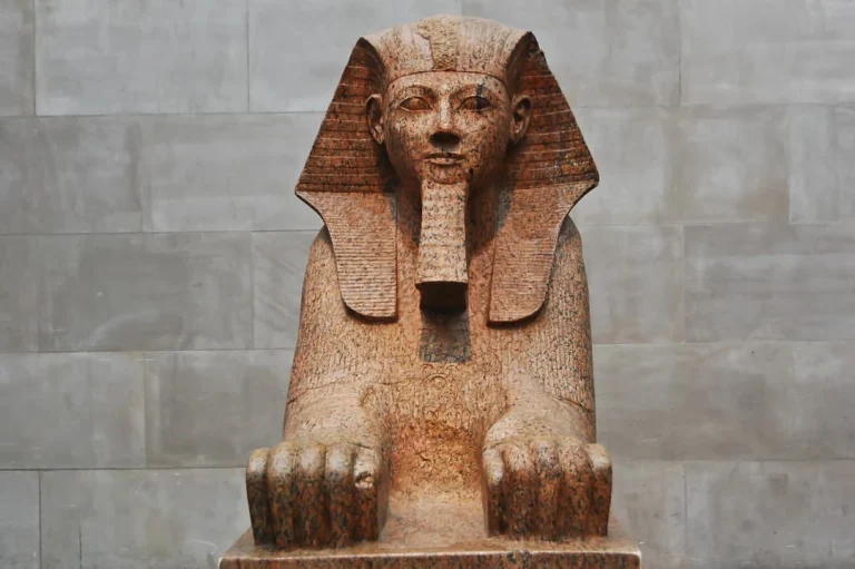 Een stenen sfinxbeeld met beschadigd gezicht tentoongesteld tegen een grijze stenen muur in het Metropolitan Museum of Art in New York
