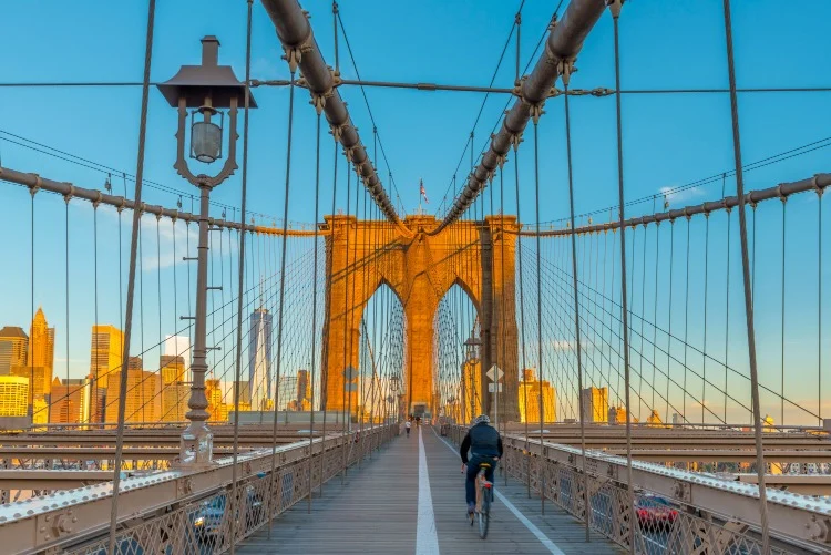 Een fietser rijdt over het houten pad van de Brooklyn Bridge in New York, met de skyline van Manhattan op de achtergrond bij zonsondergang