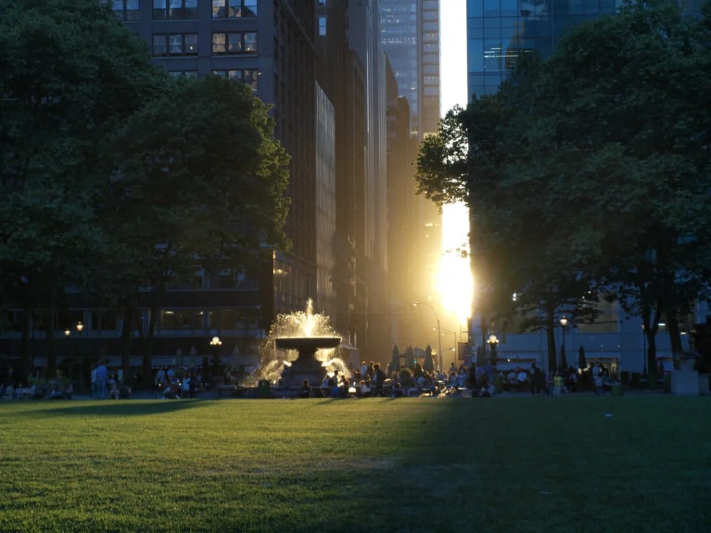 Spuitende fontein in Bryant Park bij schemering met bezoekers op het gras en fel oranje zonlicht tussen de wolkenkrabbers door