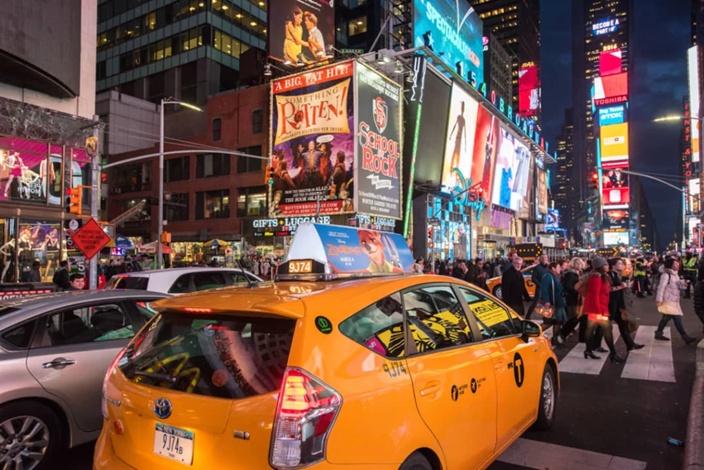 Gele taxi geparkeerd op straat op Times Square in de avond, met grote verlichte affiches van Broadway musicals erachter