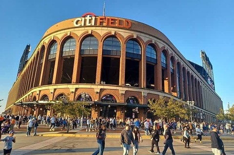 Bezoekers lopen langs de bakstenen gevel met bogen van honkbalstadion Citi Field onder een blauwe lucht