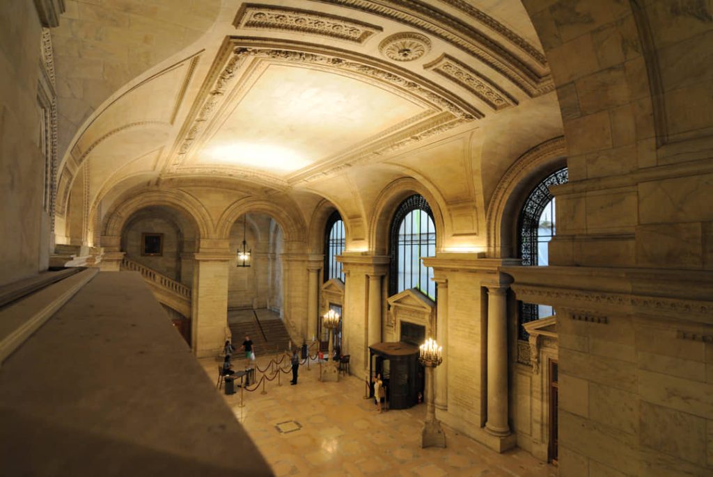 Statige gewelfde marmeren hal met bogen en een klok in de New York Public Library, bezoekers lopen op de trap