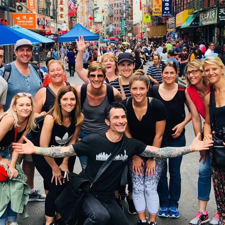 Een groep lachende toeristen poseert voor de camera op een drukke straat in Chinatown, New York City, omringd door winkels met Chinese uithangborden