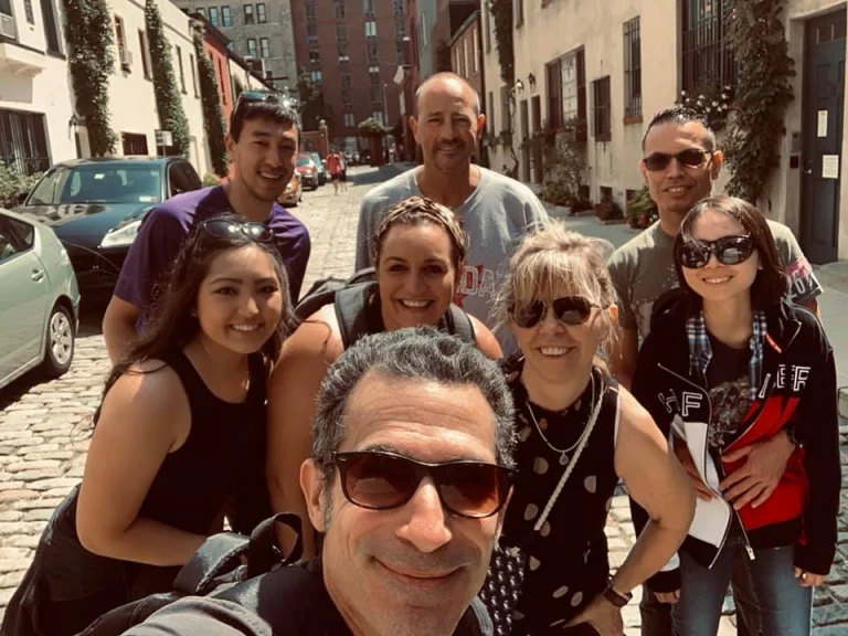 Een groep van acht lachende mensen poseert voor een selfie op een geplaveide straat tussen oude gebouwen in New York