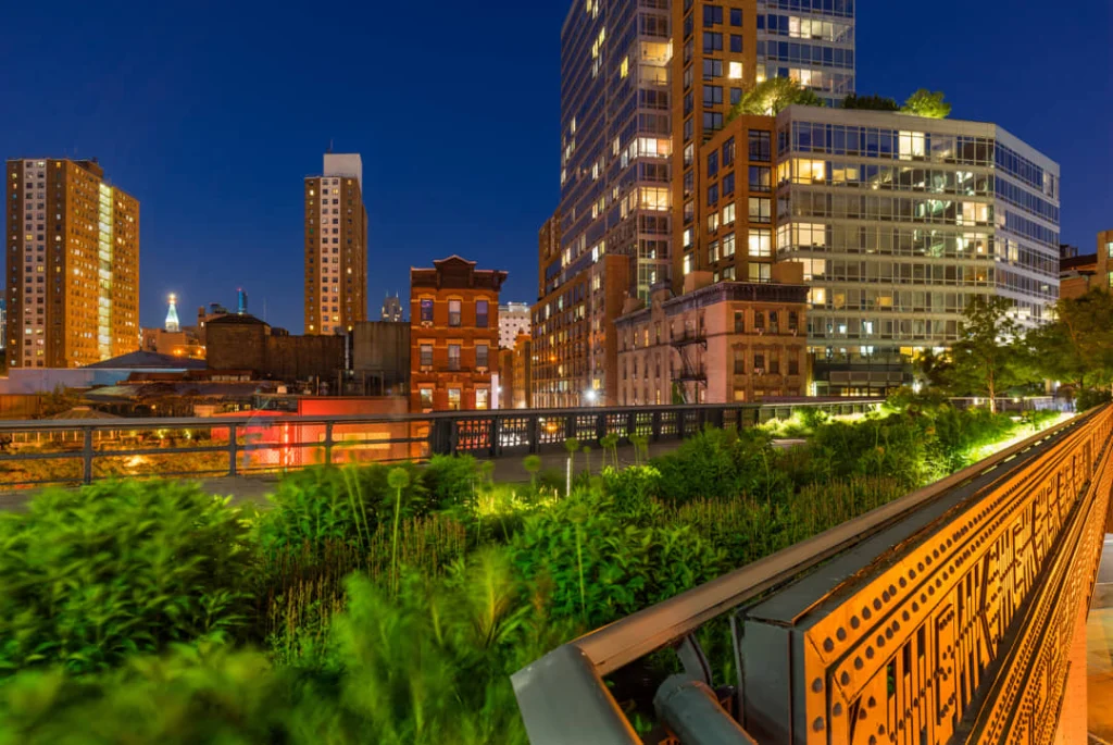 Weelderige beplanting langs de High Line in de avond met verlichte gebouwen en een spoorbrug op de achtergrond