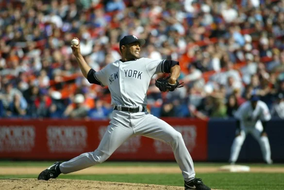 Een honkbalpitcher in een New York uniform werpt de bal richting het thuisplaat voor een volle tribune met toeschouwers