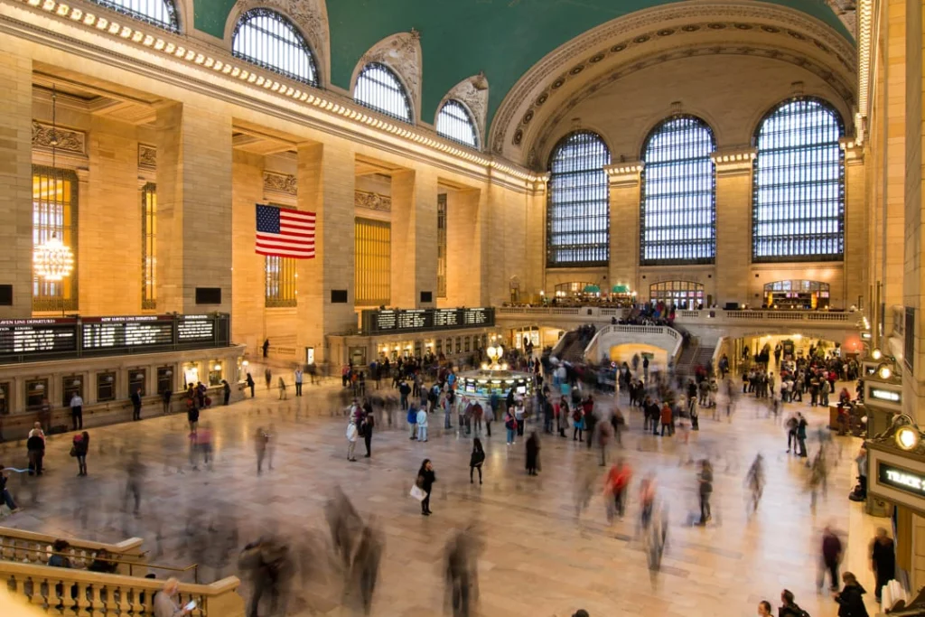 Drukke hoofdhal van Grand Central Terminal met een groen plafond, hoge ramen, een Amerikaanse vlag en talloze reizigers die zich door de ruimte bewegen