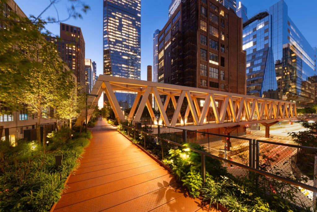 Verlicht wandelpad van de High Line onder een houten vakwerkconstructie met de skyline van gebouwen op de achtergrond bij schemering