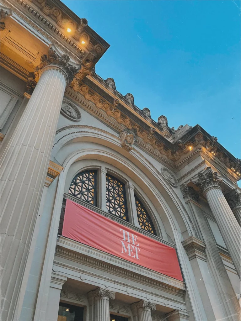 De ingang van het Metropolitan Museum of Art met een rode banier en verlichte zuilen tegen een blauwe avondlucht in New York