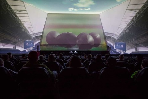 Bezoekers zitten in een donkere zaal met een groot gebogen scherm waarop American football wordt getoond