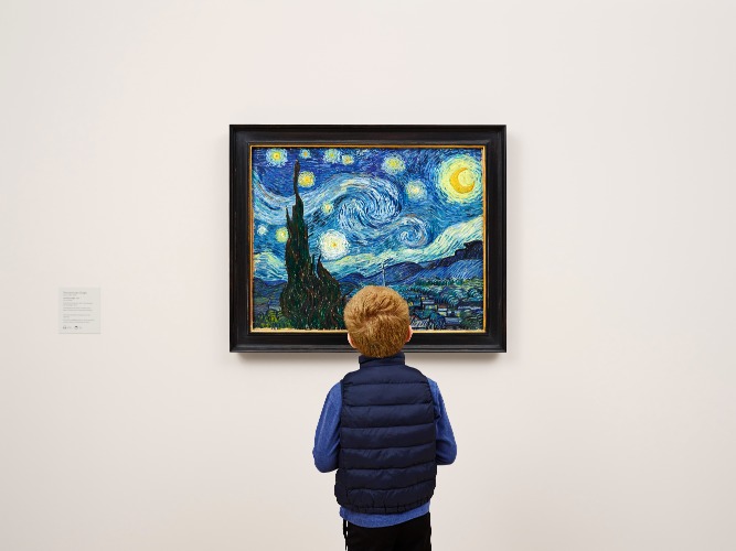 Een jongen in donkerblauw vest kijkt van achteren naar het schilderij Sterrennacht van Van Gogh aan een museumwand