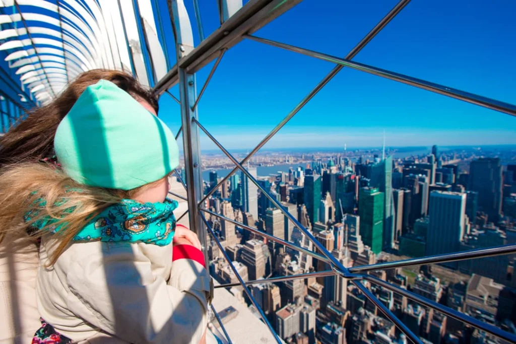 Kind met muts en sjaal kijkt door het hek van het openluchtdek van het Empire State Building over New York