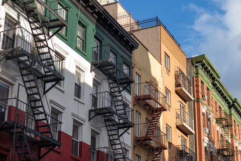 Rij appartementsgebouwen in wit, groen, geel en rood met zwarte brandtrappen tegen een blauwe lucht in Little Italy