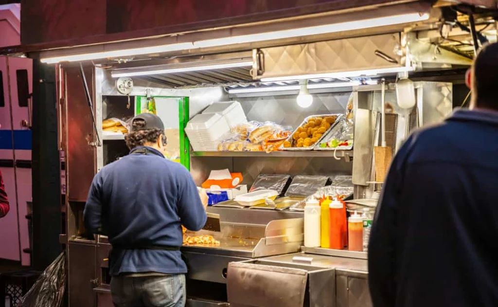 Kok met rug naar de camera bereidt eten in een verlichte foodtruck terwijl een klant op zijn bestelling wacht