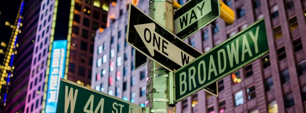 Straatnaambordjes voor Broadway en West 44th Street aan een lantaarnpaal in Times Square New York, met verlichte gebouwen op de achtergrond in de avond