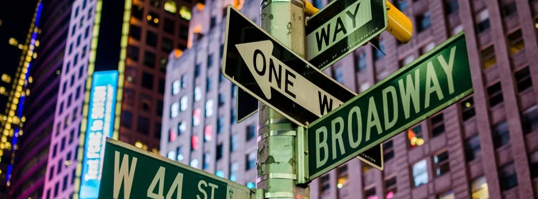 Straatnaambordjes voor Broadway en West 44th Street aan een lantaarnpaal in Times Square New York, met verlichte gebouwen op de achtergrond in de avond