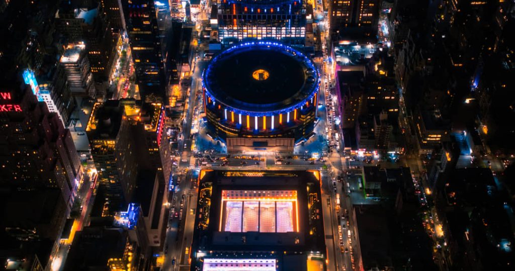 Luchtfoto bij nacht van het ronde dak van Madison Square Garden en het verlichte Pennsylvania Station ernaast tussen straten vol lichten