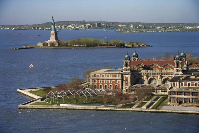 Luchtfoto van het groene Vrijheidsbeeld op zijn sokkel omringd door water met de gebouwen van Ellis Island op de achtergrond