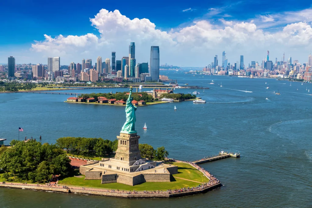Luchtfoto van het Vrijheidsbeeld op Liberty Island met de skyline van Manhattan en Jersey City in de achtergrond en blauw water van de haven eromheen