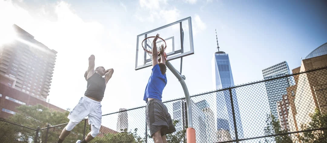 Twee mannen spelen basketbal op een omheind stadsveld in New York, met de wolkenkrabbers van Manhattan op de achtergrond