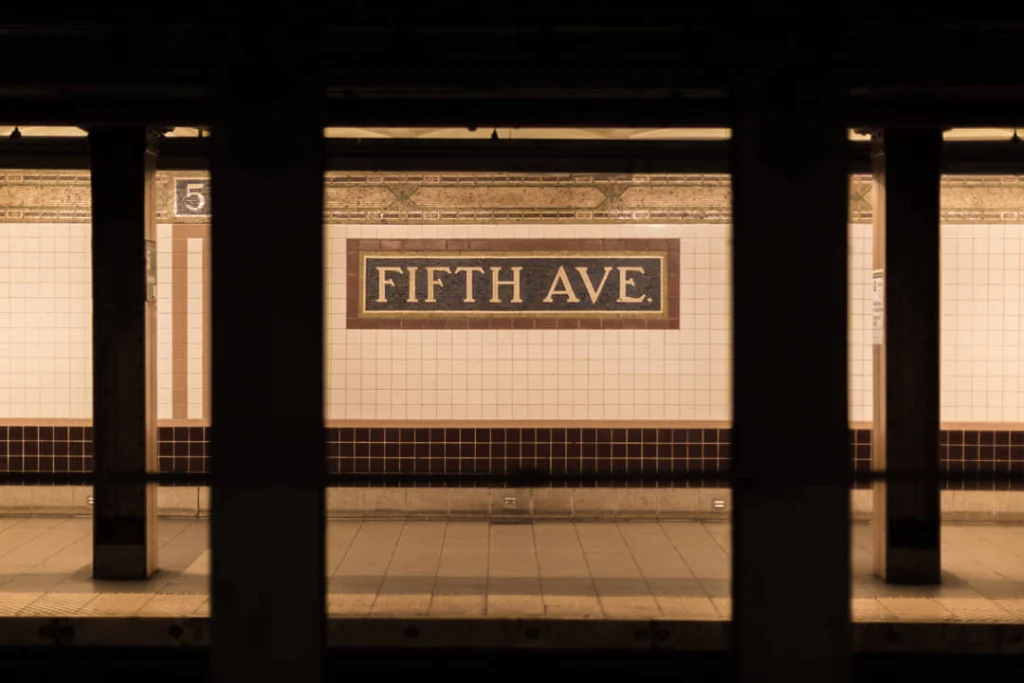 Tegelmuur in een metrostation in New York met een bord met de tekst Fifth Avenue tussen de perronzuilen