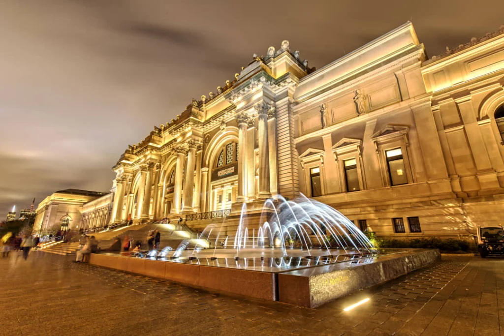 Verlichte gevel van het Metropolitan Museum of Art met spuitende fonteinen op het plein ervoor in de avond